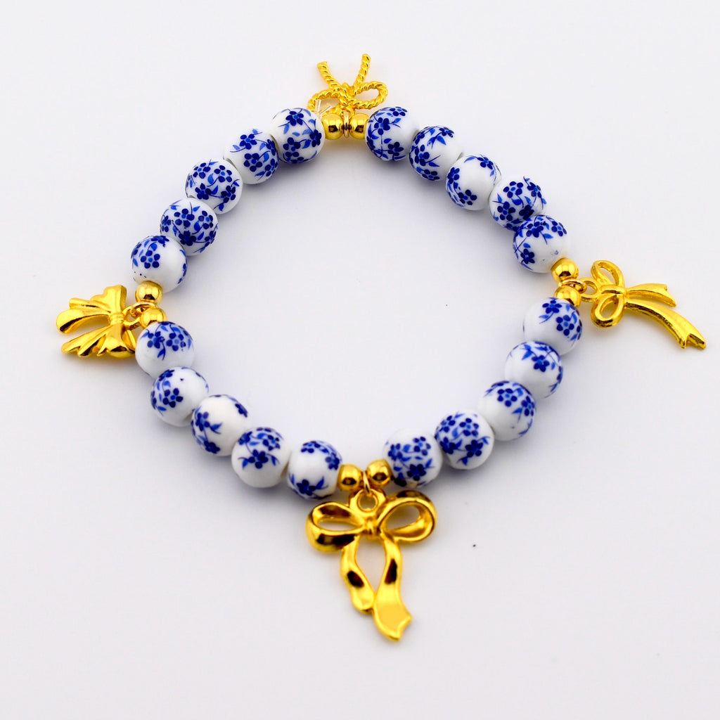 A beaded bracelet featuring round white ceramic beads with a blue floral pattern. The bracelet is accented with three gold-toned metal bow charms and small gold-toned spacer beads interspersed between the ceramic beads.
