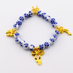 A beaded bracelet featuring round white ceramic beads with a blue floral pattern. The bracelet is accented with three gold-toned metal bow charms and small gold-toned spacer beads interspersed between the ceramic beads.