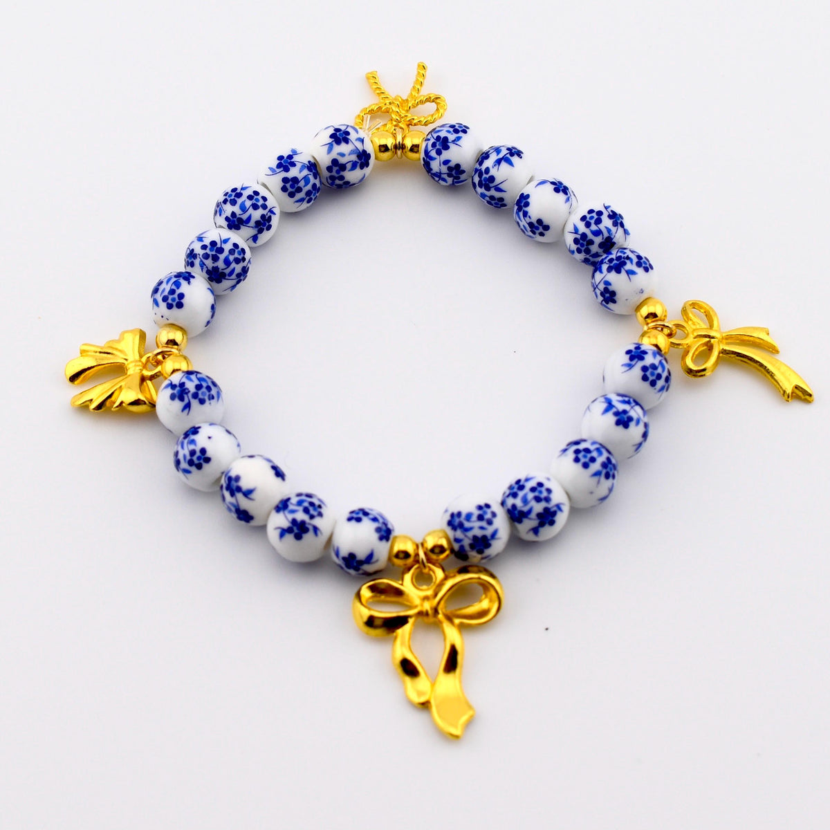 A beaded bracelet featuring round white ceramic beads with a blue floral pattern. The bracelet is accented with three gold-toned metal bow charms and small gold-toned spacer beads interspersed between the ceramic beads.