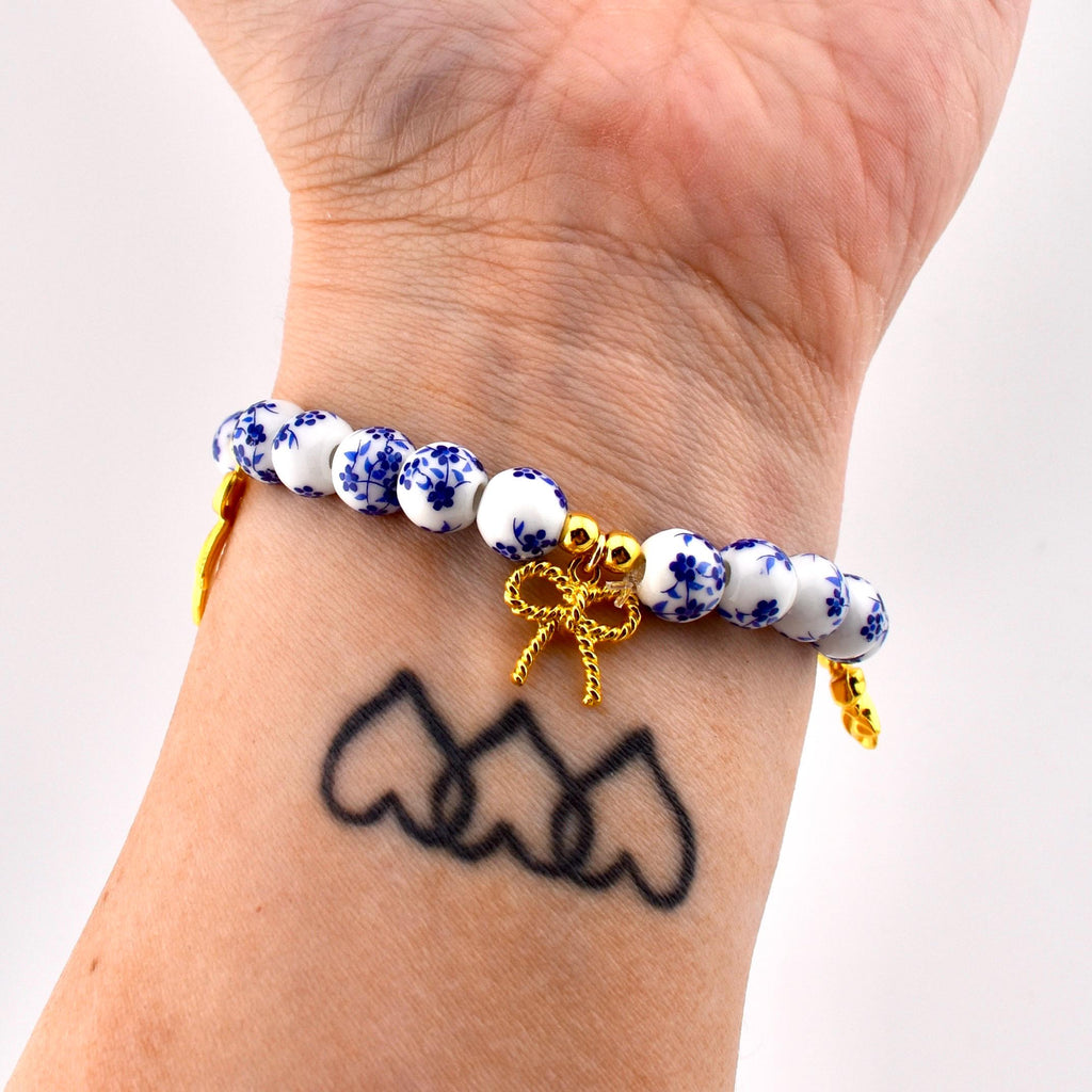A beaded bracelet featuring round white ceramic beads with a blue floral pattern. The bracelet includes gold-toned metal spacers and a central gold-toned bow charm. The beads are strung on an elastic cord, creating a circular design.