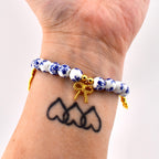 A beaded bracelet featuring round white ceramic beads with a blue floral pattern. The bracelet includes gold-toned metal spacers and a central gold-toned bow charm. The beads are strung on an elastic cord, creating a circular design.