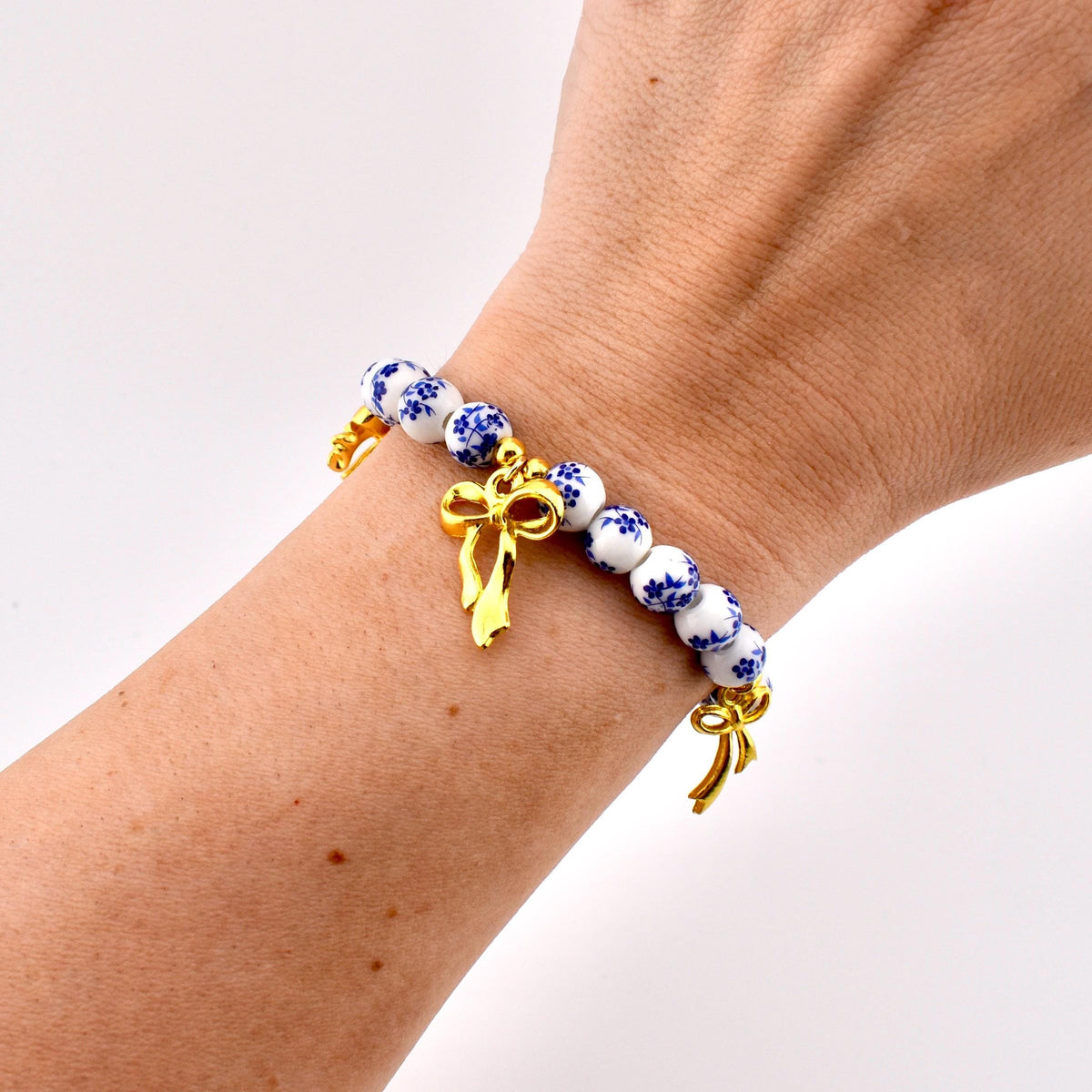 A beaded bracelet featuring round white ceramic beads with a blue floral pattern. The bracelet is accented with two polished gold-tone metal bow charms hanging from the strand, creating a decorative contrast against the white and blue beads.