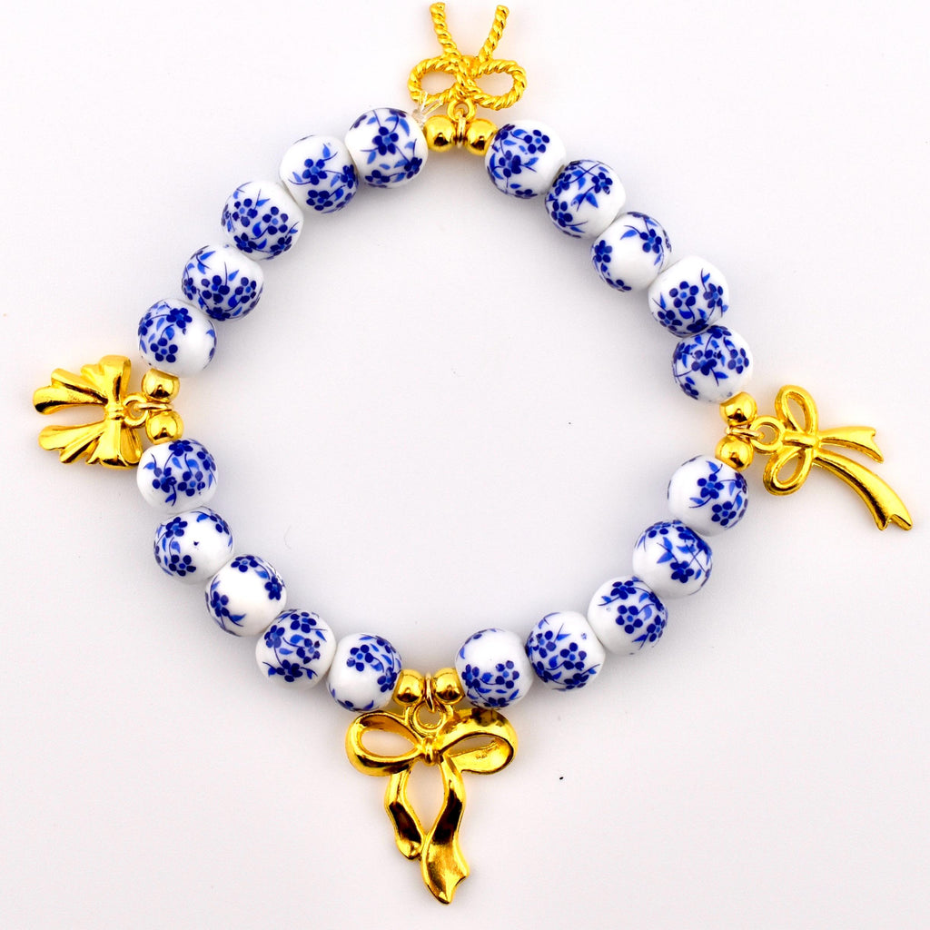 A circular bracelet featuring white spherical beads decorated with a blue floral pattern. The bracelet is accented with four gold-toned metal bow charms spaced evenly around the circumference, connected by small gold-toned metal beads.