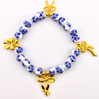 A circular bracelet featuring white spherical beads decorated with a blue floral pattern. The bracelet is accented with four gold-toned metal bow charms spaced evenly around the circumference, connected by small gold-toned metal beads.