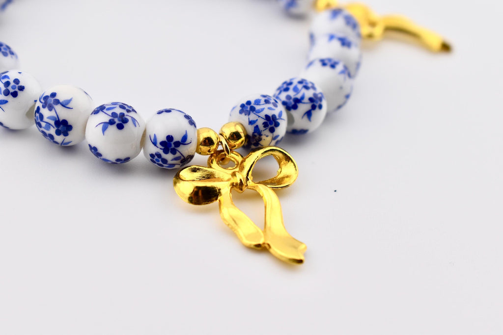 A bracelet featuring round white ceramic beads decorated with a blue floral pattern. The bracelet includes a polished gold-toned metal bow charm suspended from a small gold-toned bead, creating a contrast between the white, blue, and gold.