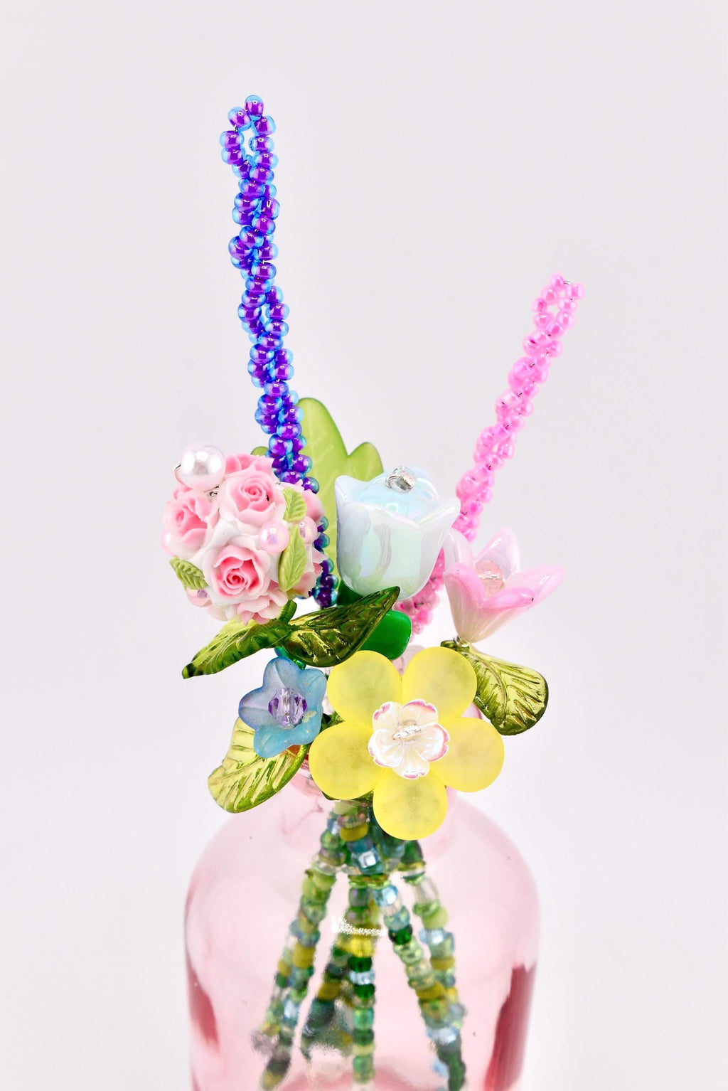 A decorative bouquet of artificial flowers crafted from beads and resin, featuring pink roses, a blue tulip, a yellow blossom, and green leaves. The stems are made of green beads, and the arrangement is placed inside a small pink glass bottle.