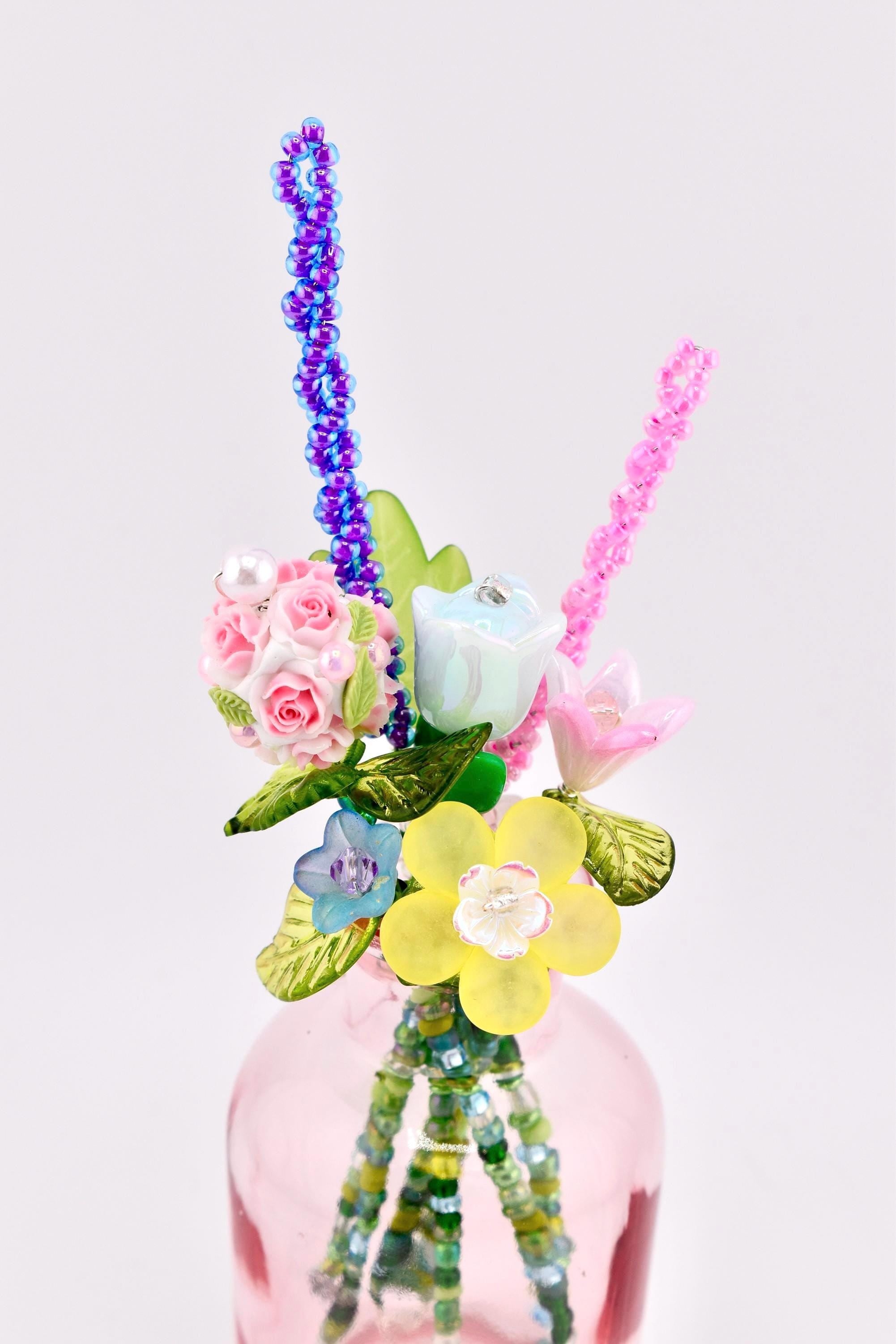 A decorative bouquet of artificial flowers crafted from beads and resin, featuring pink roses, a blue tulip, a yellow blossom, and green leaves. The stems are made of green beads, and the arrangement is placed inside a small pink glass bottle.