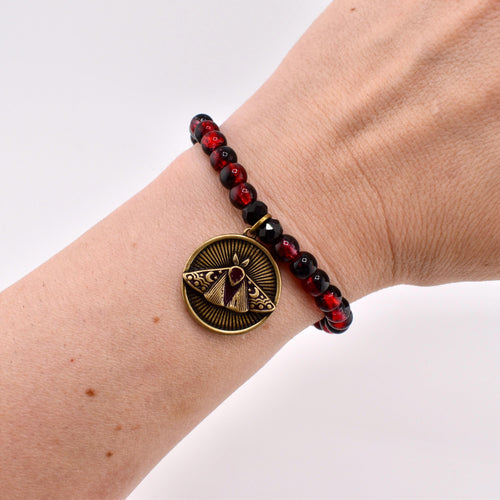 A beaded bracelet featuring alternating dark red and black spherical beads. A circular bronze-toned metal charm hangs from the bracelet, displaying an embossed moth design with sunburst rays and small crescent moon details in the background.