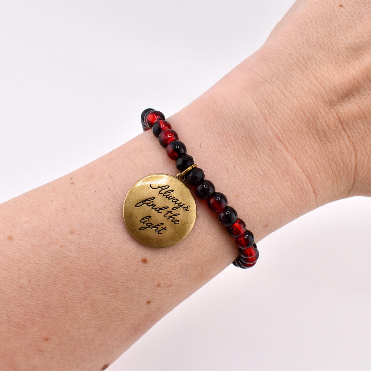 A beaded bracelet featuring alternating black and translucent red spherical beads. A round, antique brass-colored metal charm hangs from the bracelet, engraved with the black cursive text Always find the light.