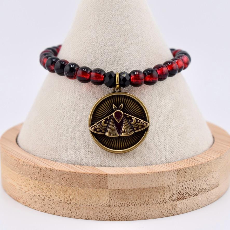 A beaded bracelet featuring translucent red and black spherical beads with a circular bronze-toned pendant. The pendant displays an engraved moth design with sunburst rays and crescent moon details, centered with a small red stone.