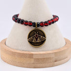 A beaded bracelet featuring translucent red and black spherical beads with a circular bronze-toned pendant. The pendant displays an engraved moth design with sunburst rays and crescent moon details, centered with a small red stone.