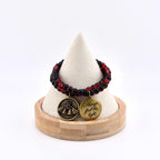 A beaded bracelet featuring alternating black and translucent red beads. Two circular brass charms hang from the bracelet: one displays an embossed moth design, and the other is engraved with the text Always find the light.
