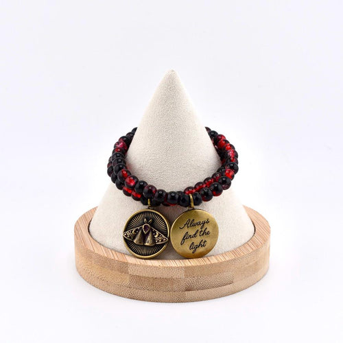 A beaded bracelet featuring alternating black and translucent red beads. Two circular brass charms hang from the bracelet: one displays an embossed moth design, and the other is engraved with the text Always find the light.
