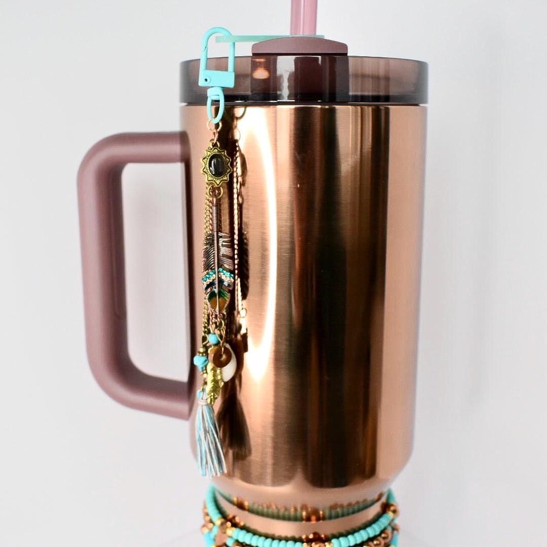 A copper-colored insulated tumbler with a handle, a teal clip, and a decorative charm. The charm features feathers, beads, and a tassel. A turquoise beaded bracelet is wrapped around the base of the tumbler. A pink straw is inserted into the lid.