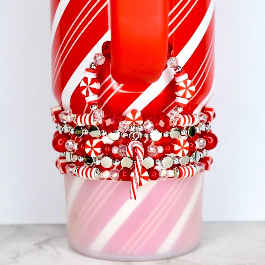 A red and white striped mug with a stack of red, white, and silver beaded bracelets. The bracelets feature candy cane charms and snowflake-shaped beads. The mug has a red handle and a pink and white striped base.