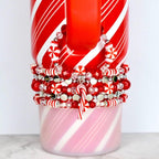 A red and white striped mug with a stack of red, white, and silver beaded bracelets. The bracelets feature candy cane charms and snowflake-shaped beads. The mug has a red handle and a pink and white striped base.