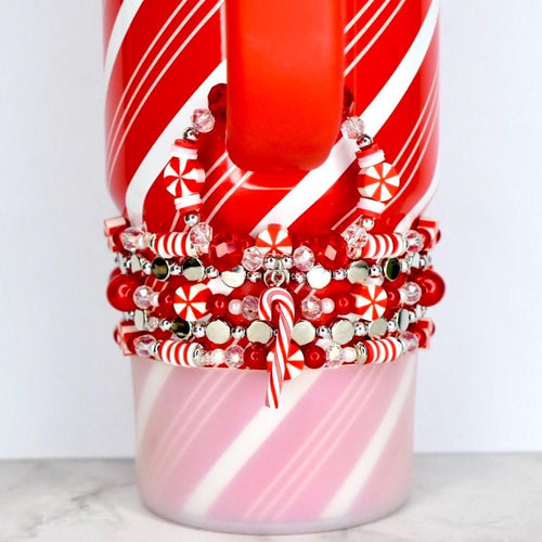 A red and white striped mug with a stack of red, white, and silver beaded bracelets. The bracelets feature candy cane charms and snowflake-shaped beads. The mug has a red handle and a pink and white striped base.