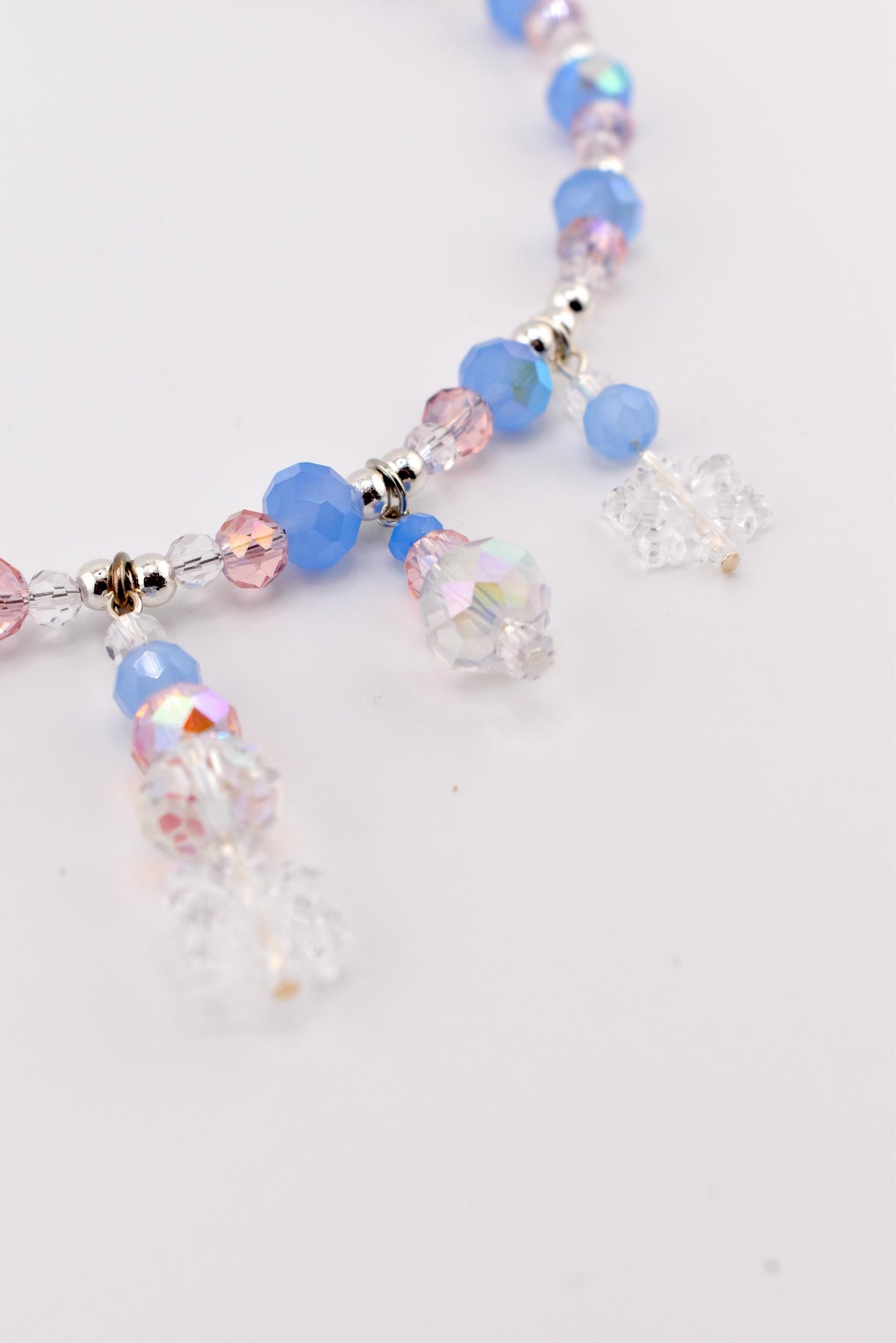 A beaded necklace featuring a strand of alternating light blue and pink faceted beads interspersed with small silver-toned spacers. Several dangling charms made of clear, iridescent, and blue beads hang from the main chain.