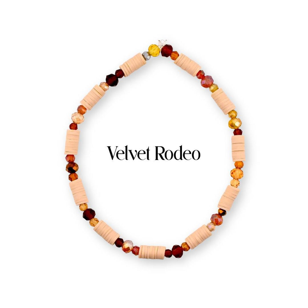 A circular beaded bracelet featuring alternating light peach cylindrical clay beads and faceted glass beads in shades of deep red, amber, and gold. The text Velvet Rodeo is centered within the bracelet loop in black lettering.