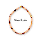 A circular beaded bracelet featuring alternating light peach cylindrical clay beads and faceted glass beads in shades of deep red, amber, and gold. The text Velvet Rodeo is centered within the bracelet loop in black lettering.
