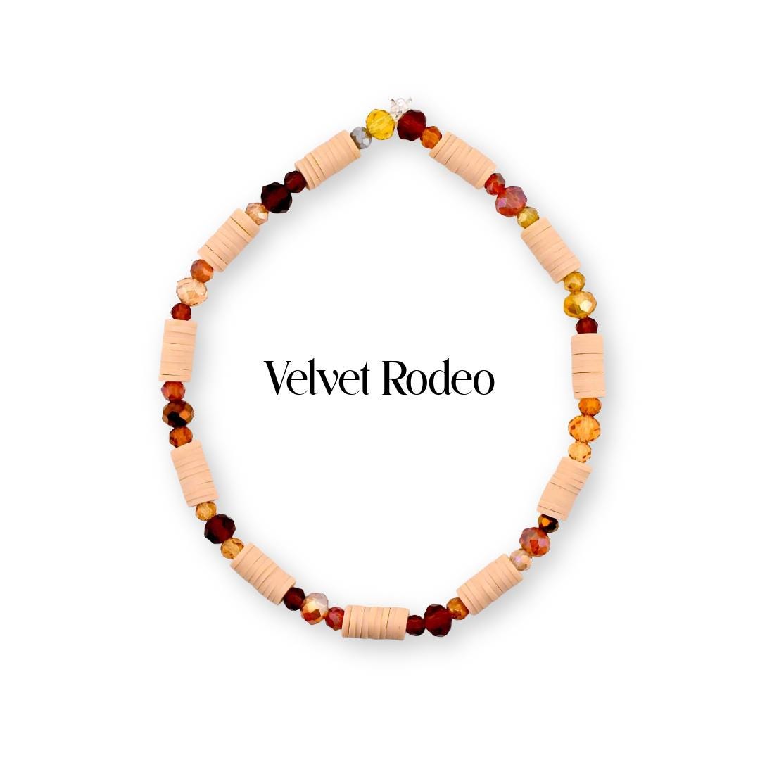 A circular beaded bracelet featuring alternating light peach cylindrical clay beads and faceted glass beads in shades of deep red, amber, and gold. The text Velvet Rodeo is centered within the bracelet loop in black lettering.
