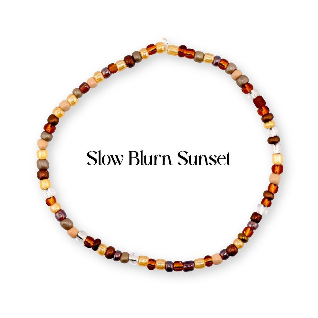 A circular beaded bracelet featuring a variety of small glass beads in shades of brown, tan, gold, and clear. The text "Slow Blurn Sunset" is printed in the center of the circular arrangement of beads against a plain white background.