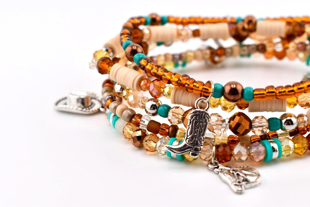 A multi-strand beaded bracelet featuring amber, brown, and turquoise glass beads, beige disc spacers, and silver-toned metal charms shaped like a cowboy hat, a cowboy boot, and a horse head, all strung on a flexible elastic cord.