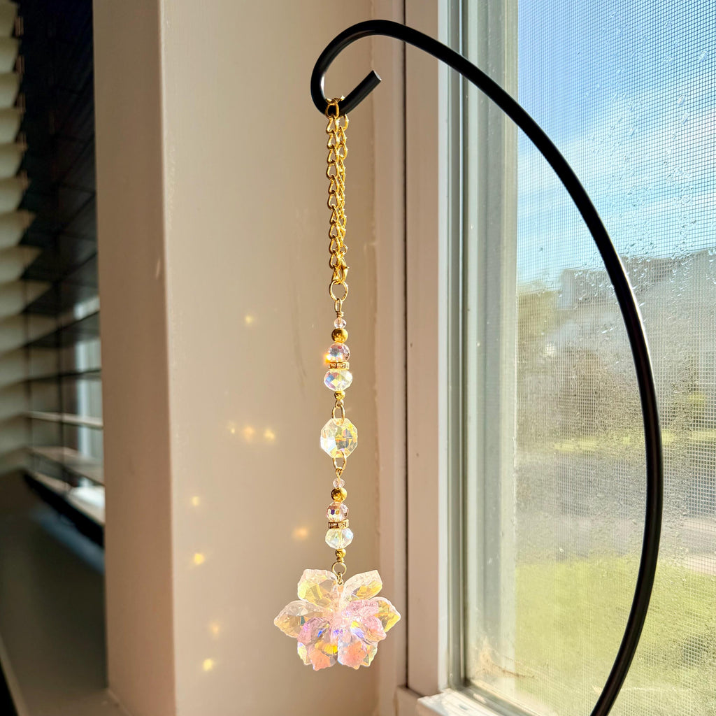 A hanging suncatcher ornament featuring a gold-toned metal chain connected to a series of clear, iridescent, multifaceted glass beads and a central flower-shaped crystal pendant that reflects light with a soft, prismatic glow.