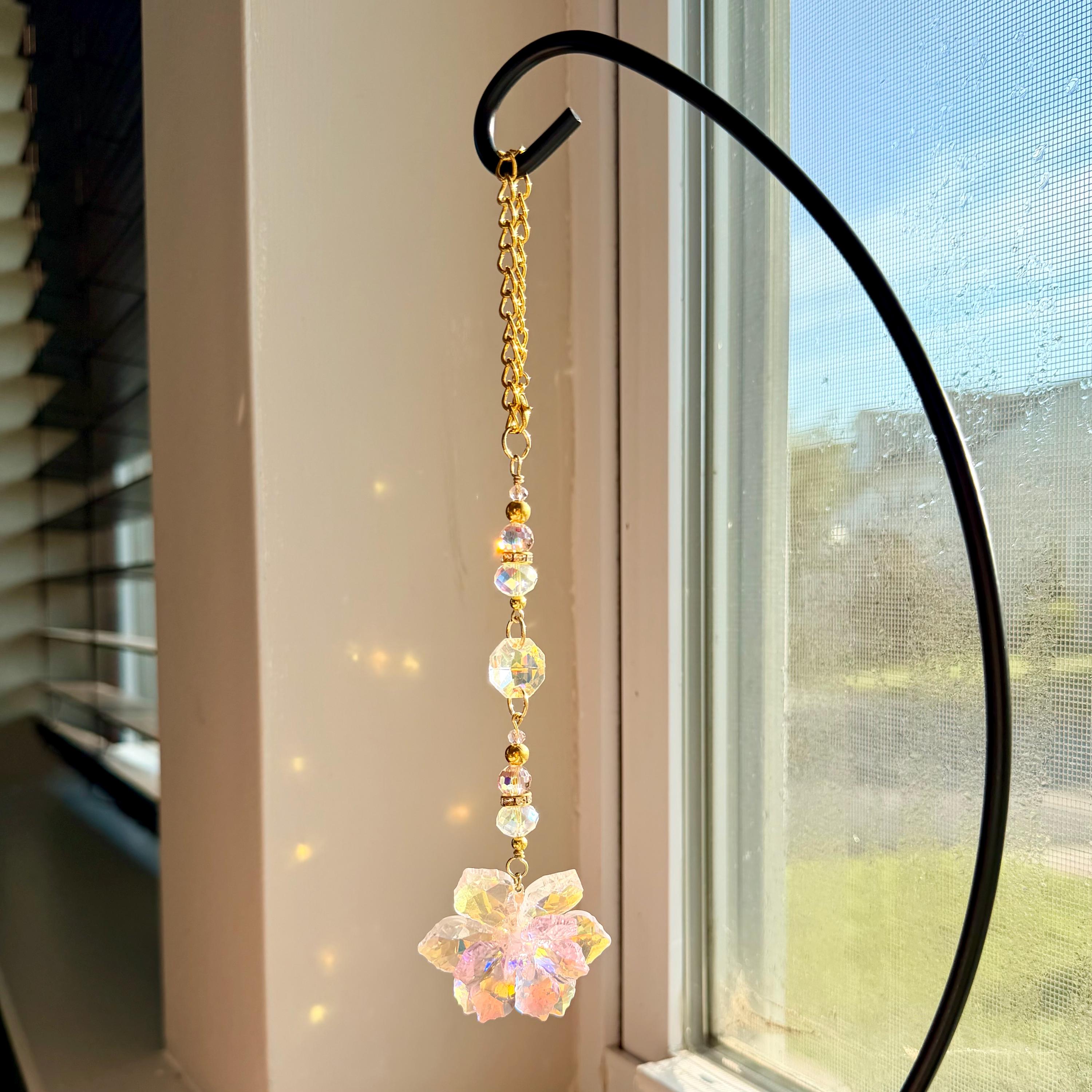 A hanging suncatcher ornament featuring a gold-toned metal chain connected to a series of clear, iridescent, multifaceted glass beads and a central flower-shaped crystal pendant that reflects light with a soft, prismatic glow.