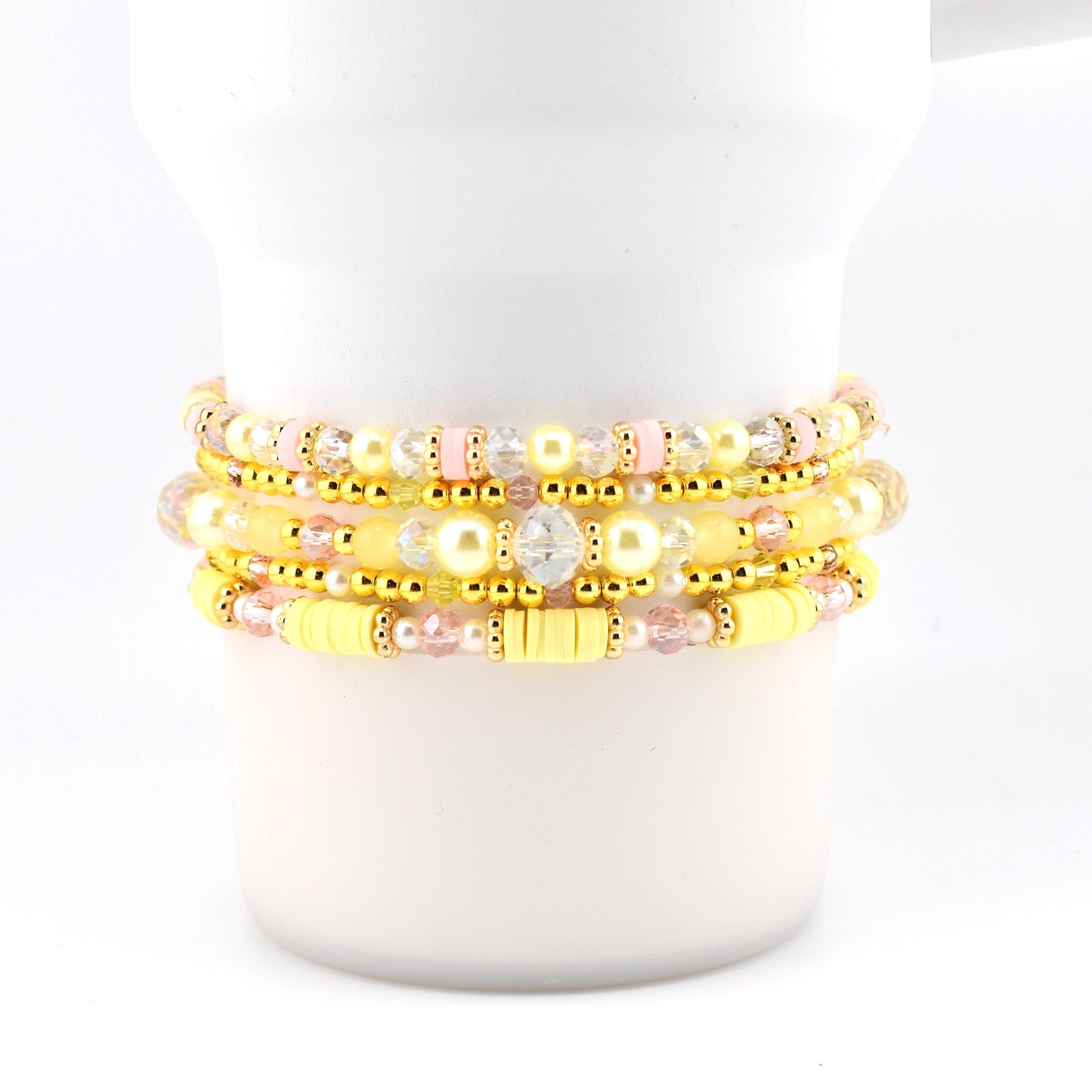 A stack of four beaded bracelets featuring a mix of yellow, pale pink, and clear crystal beads, interspersed with metallic gold-tone spacer beads and flat yellow polymer clay discs, arranged in a layered design on a white cylindrical form.