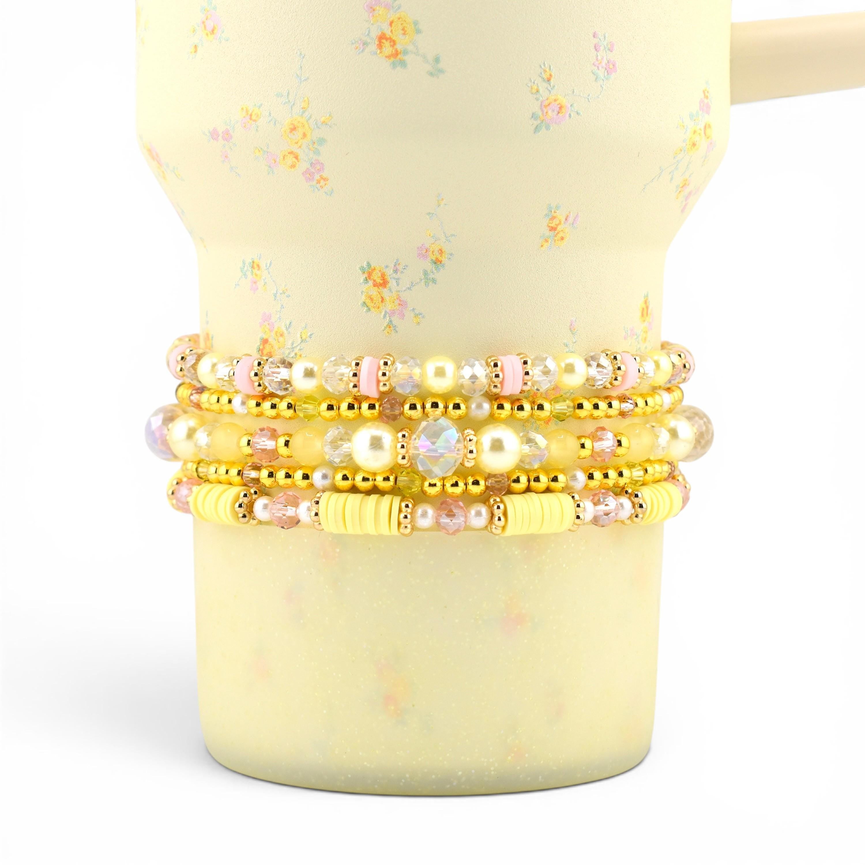 A stack of five beaded bracelets featuring a mix of yellow, pink, and white beads. The design includes gold-toned metallic spacers, clear faceted crystals, smooth round pearls, and cylindrical yellow polymer clay beads.