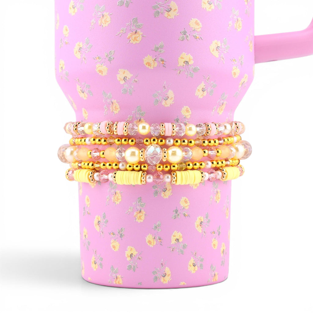A stack of four beaded bracelets featuring a mix of gold-tone metallic beads, clear faceted crystals, pale yellow clay discs, and cream-colored faux pearls, wrapped around a pink tumbler with a yellow and green floral pattern.