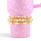 A stack of four beaded bracelets featuring a mix of gold-tone metallic beads, clear faceted crystals, pale yellow clay discs, and cream-colored faux pearls, wrapped around a pink tumbler with a yellow and green floral pattern.