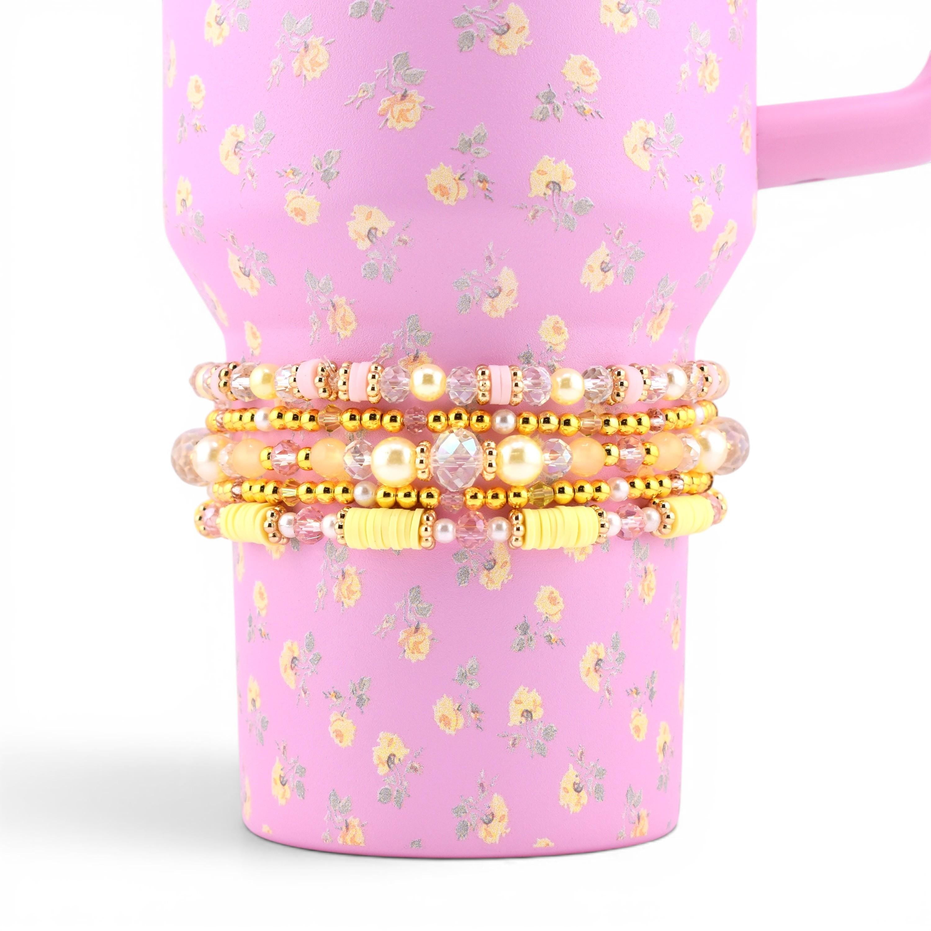 A stack of four beaded bracelets featuring a mix of gold-tone metallic beads, clear faceted crystals, pale yellow clay discs, and cream-colored faux pearls, wrapped around a pink tumbler with a yellow and green floral pattern.