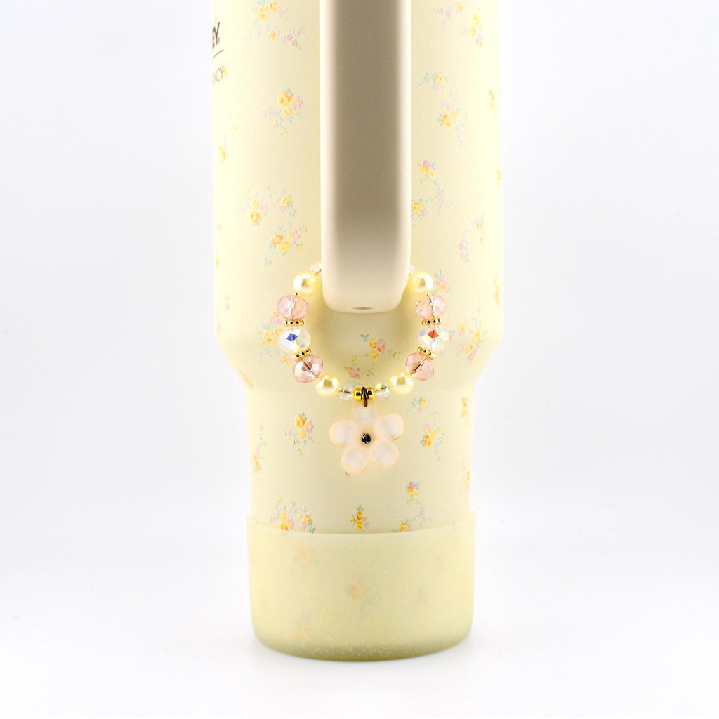 A pale yellow tumbler with a small floral pattern features a beaded charm attached to its handle. The circular charm consists of alternating cream-colored pearls, translucent pink faceted beads, and a single light pink flower pendant.