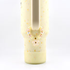 A pale yellow tumbler with a small floral pattern features a beaded charm attached to its handle. The circular charm consists of alternating cream-colored pearls, translucent pink faceted beads, and a single light pink flower pendant.