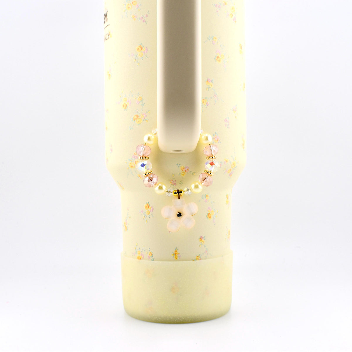 A pale yellow tumbler with a small floral pattern features a beaded charm attached to its handle. The circular charm consists of alternating cream-colored pearls, translucent pink faceted beads, and a single light pink flower pendant.