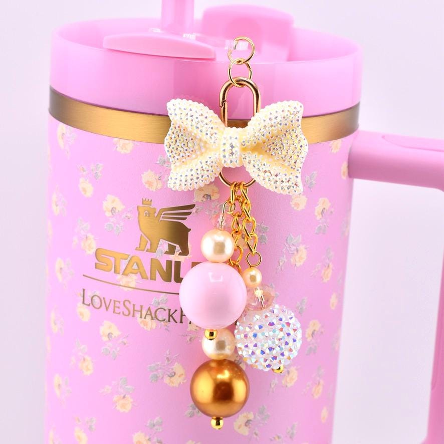 A pink tumbler with a floral pattern and the text STANLEY LOVESHACKFANCY. Attached to the lid is a gold-toned charm featuring a glittery cream bow, a large pink bead, a metallic gold bead, cream pearls, and a sparkling crystal sphere.