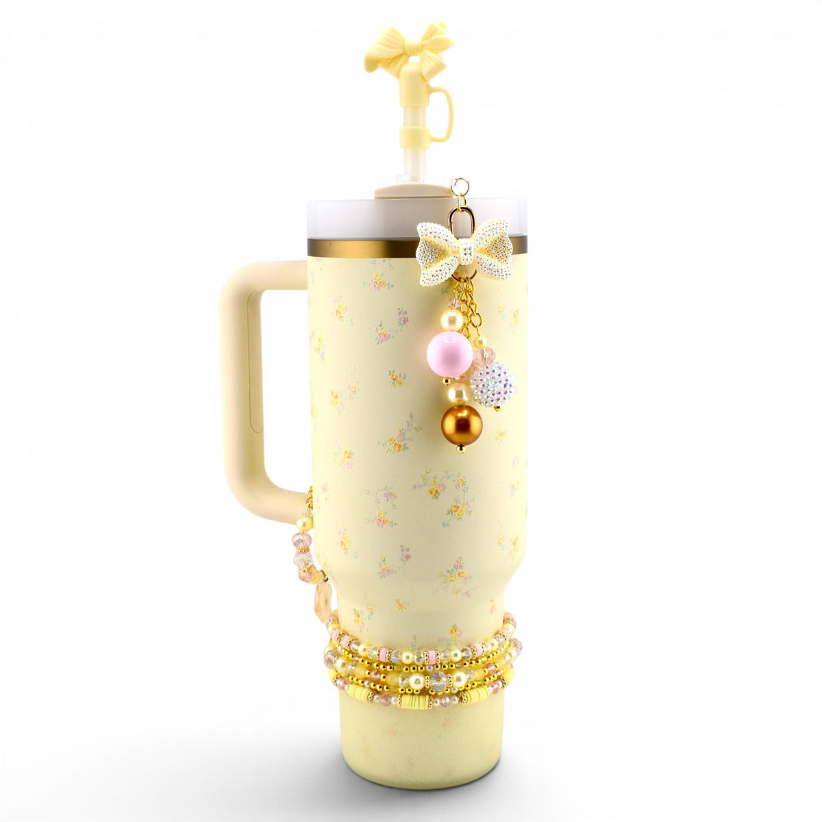 A pale yellow tumbler with a handle and a straw topper shaped like a bow. The tumbler features a small floral pattern and is decorated with a dangling charm featuring a bow, pink and gold beads, and a beaded bracelet around the base.