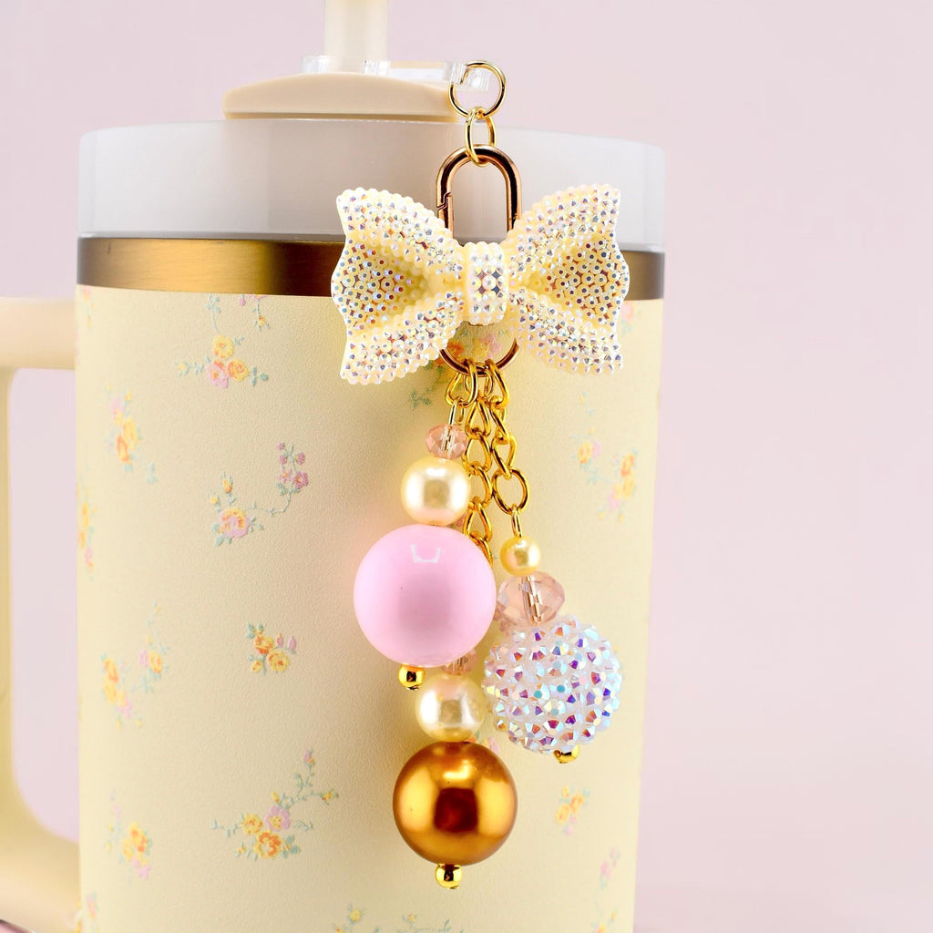 A decorative charm featuring a glittery cream-colored bow attached to a gold-tone metal ring. The charm includes a cluster of beads in various sizes and finishes, including matte pink, metallic gold, cream, and iridescent crystal spheres.