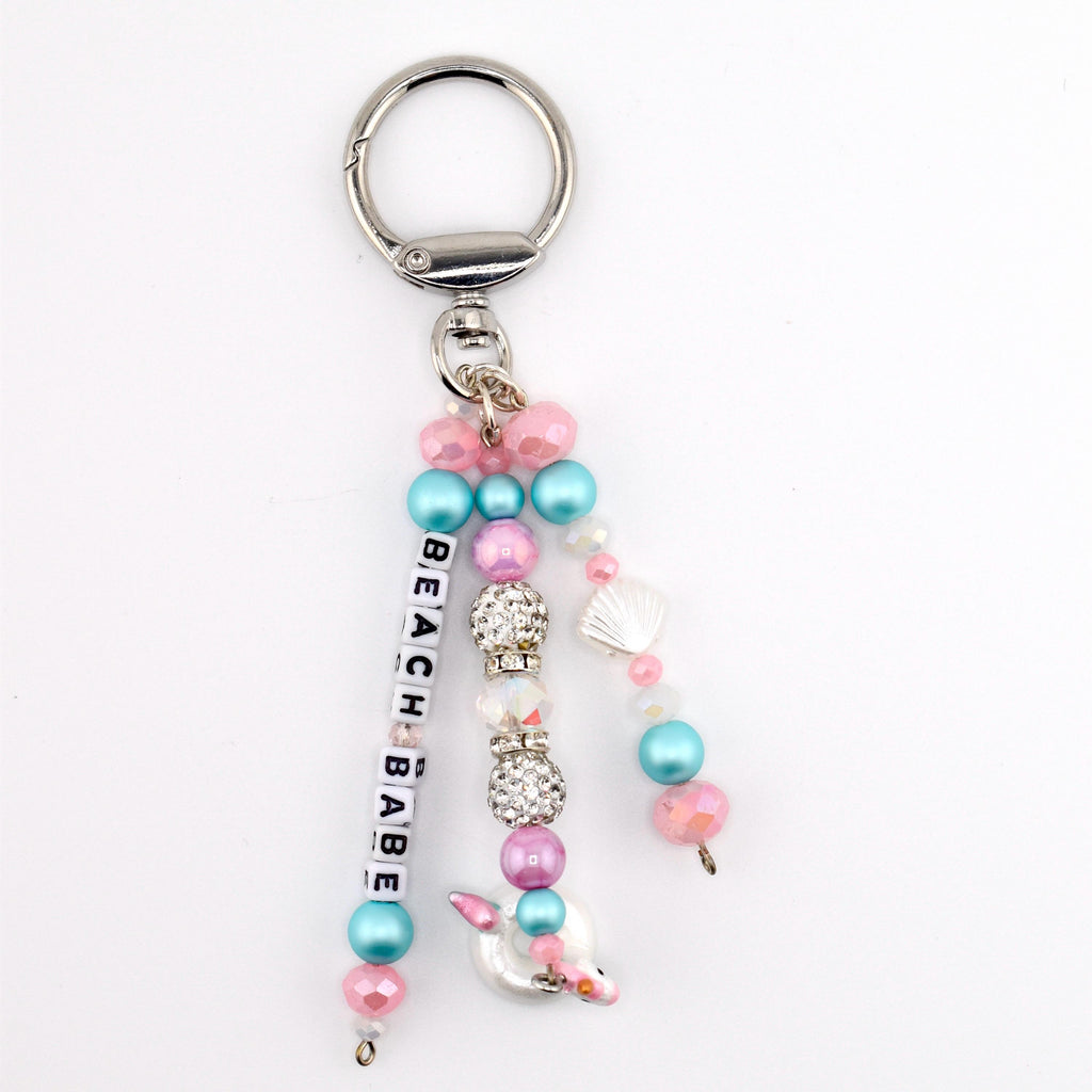 A silver-toned metal keychain featuring three dangling strands of beads. One strand displays white square letter beads spelling BEACH BABE. The design includes pink, blue, and clear crystal-encrusted beads, plus a small white seashell charm.