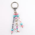 A silver-toned metal keychain featuring three dangling strands of beads. One strand displays white square letter beads spelling BEACH BABE. The design includes pink, blue, and clear crystal-encrusted beads, plus a small white seashell charm.