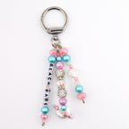 A silver-toned metal keychain featuring three dangling strands of beads. One strand displays white square letter beads spelling BEACH BABE. The design includes pink, blue, and clear crystal-encrusted beads, plus a small white seashell charm.