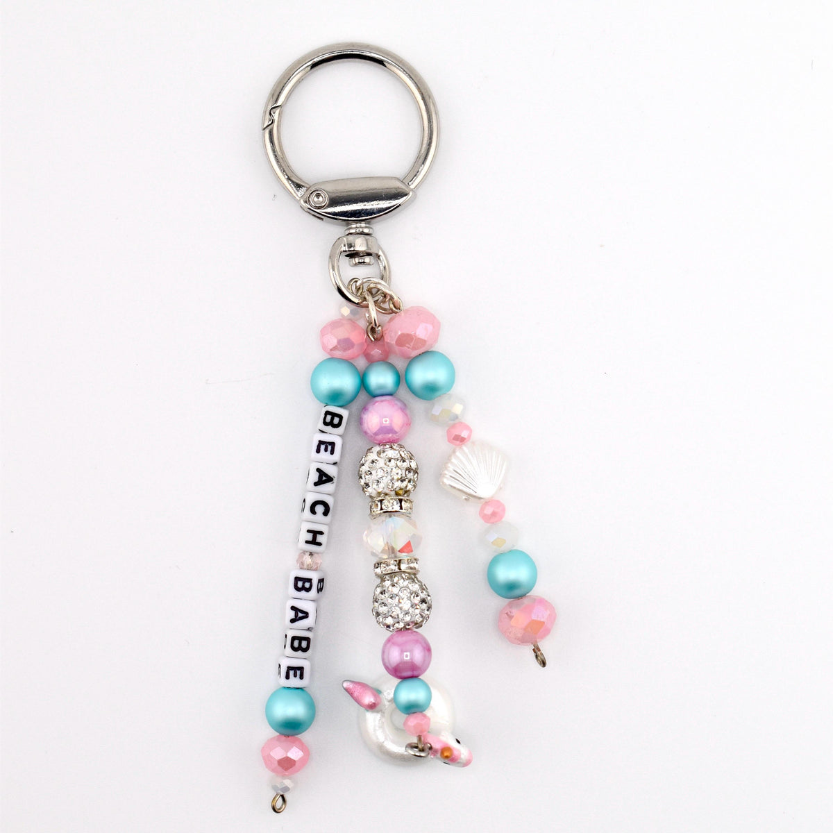 A silver-toned metal keychain featuring three dangling strands of beads. One strand displays white square letter beads spelling BEACH BABE. The design includes pink, blue, and clear crystal-encrusted beads, plus a small white seashell charm.