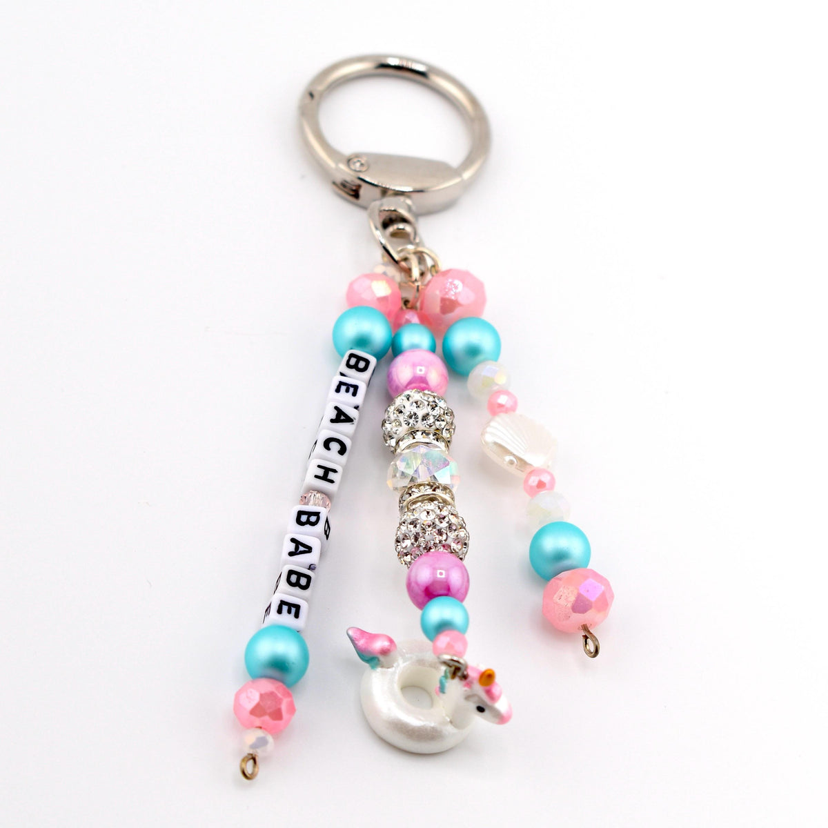 A keychain featuring a silver metal ring attached to three beaded strands. One strand includes white letter beads spelling BEACH BABE, pink and blue beads. Another has pink, blue, and rhinestone beads. The third has a unicorn float charm.