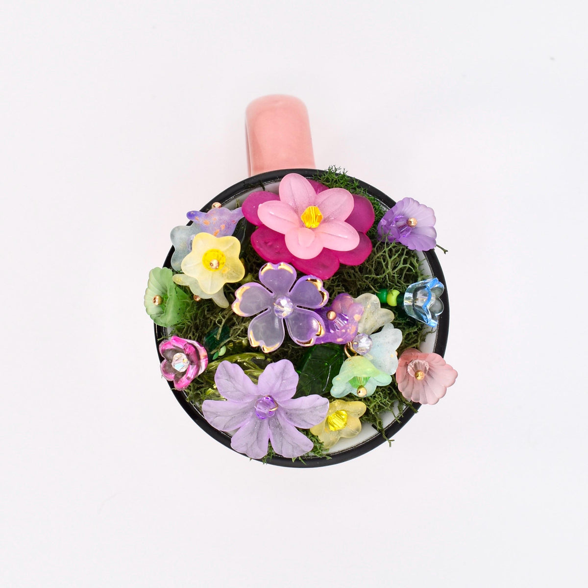 A round mug with a black rim and a pink handle, filled with green moss and an assortment of translucent, multi-colored artificial flowers in shades of pink, purple, yellow, blue, and green, each featuring a small central bead detail.