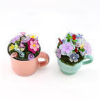 Two small ceramic mugs, one pastel pink and one light mint green, each filled with green moss and an arrangement of colorful translucent flower-shaped beads in shades of purple, pink, blue, yellow, and red with gold accents.