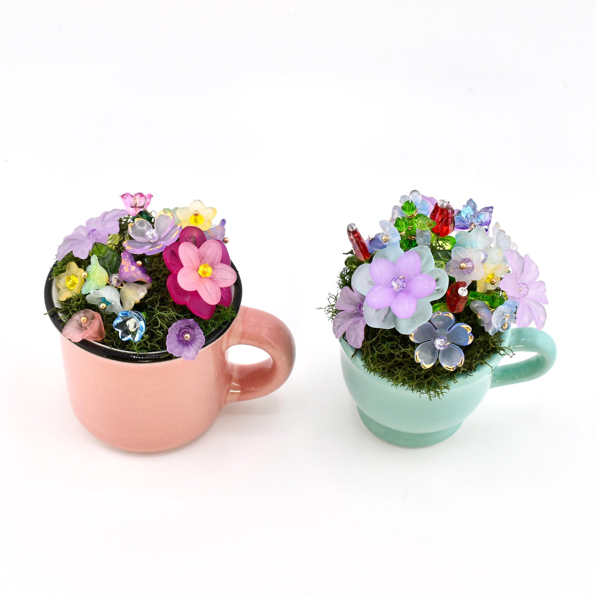 Two small ceramic mugs, one pastel pink and one light mint green, each filled with green moss and an arrangement of colorful translucent flower-shaped beads in shades of purple, pink, blue, yellow, and red with gold accents.
