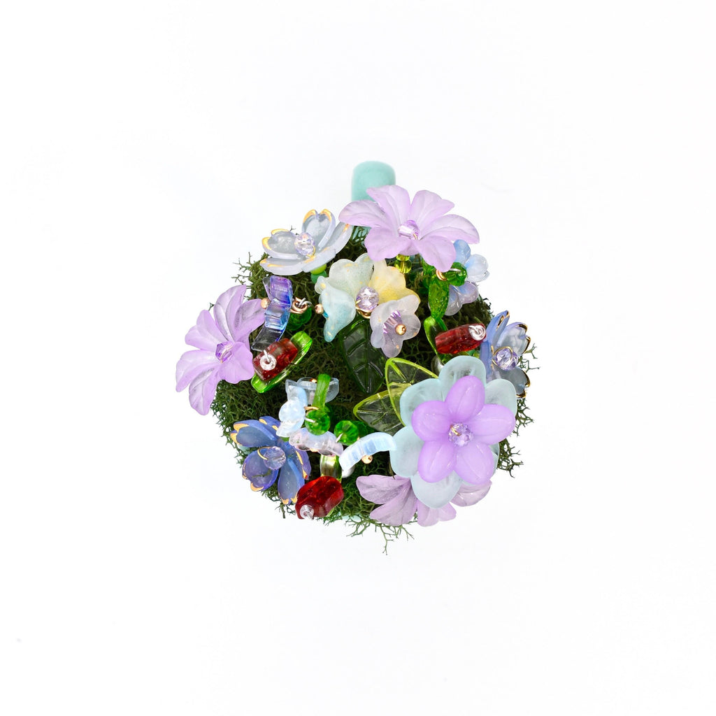 A circular decorative ornament featuring a dense cluster of artificial flowers in shades of lavender, light blue, and white, interspersed with green moss, small translucent glass beads, and tiny red accents.
