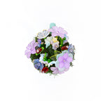 A circular decorative ornament featuring a dense cluster of artificial flowers in shades of lavender, light blue, and white, interspersed with green moss, small translucent glass beads, and tiny red accents.